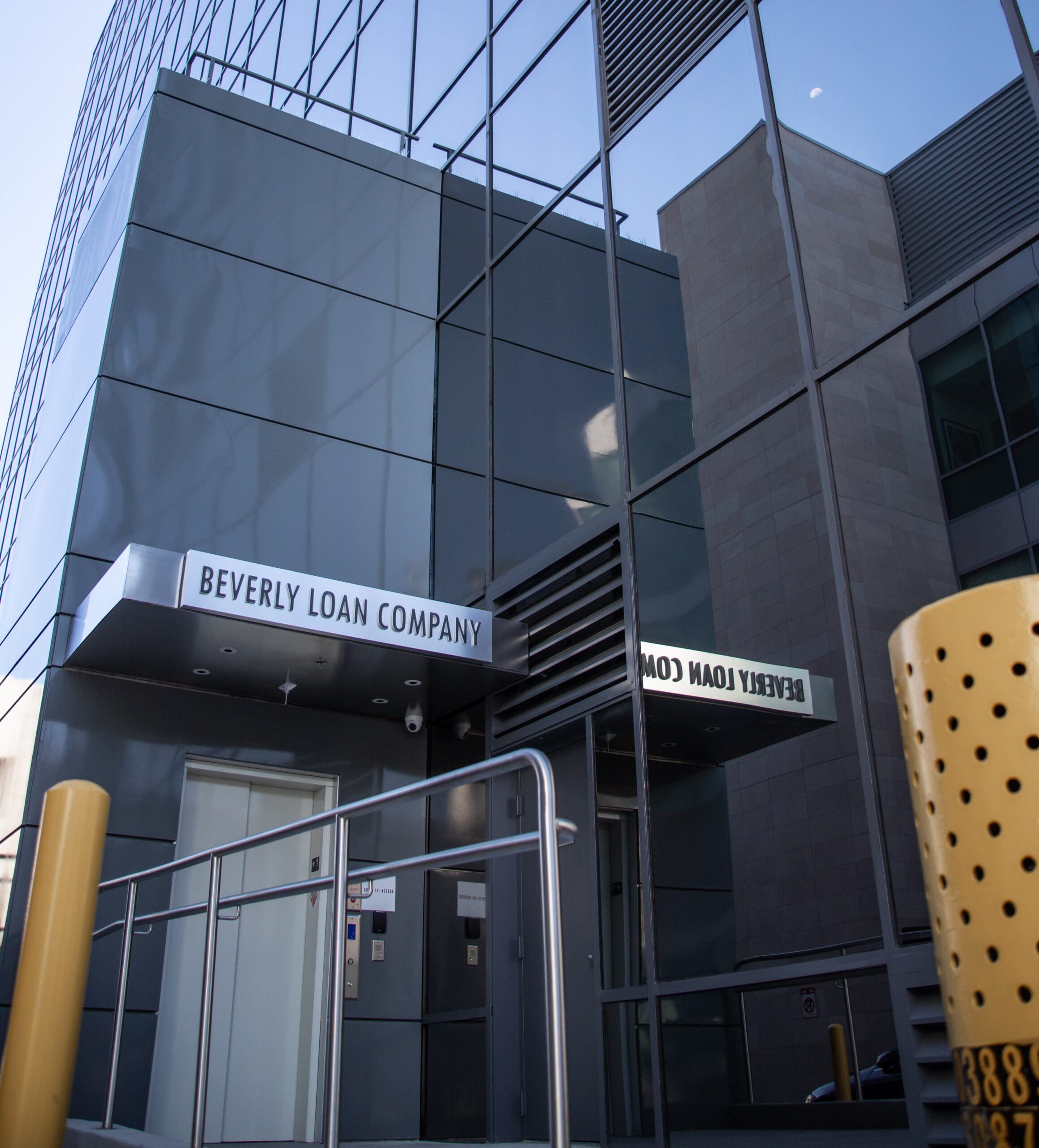 Exterior of Beverly Loan Company building with a ramp and elevator, reflecting the sky and nearby structures.