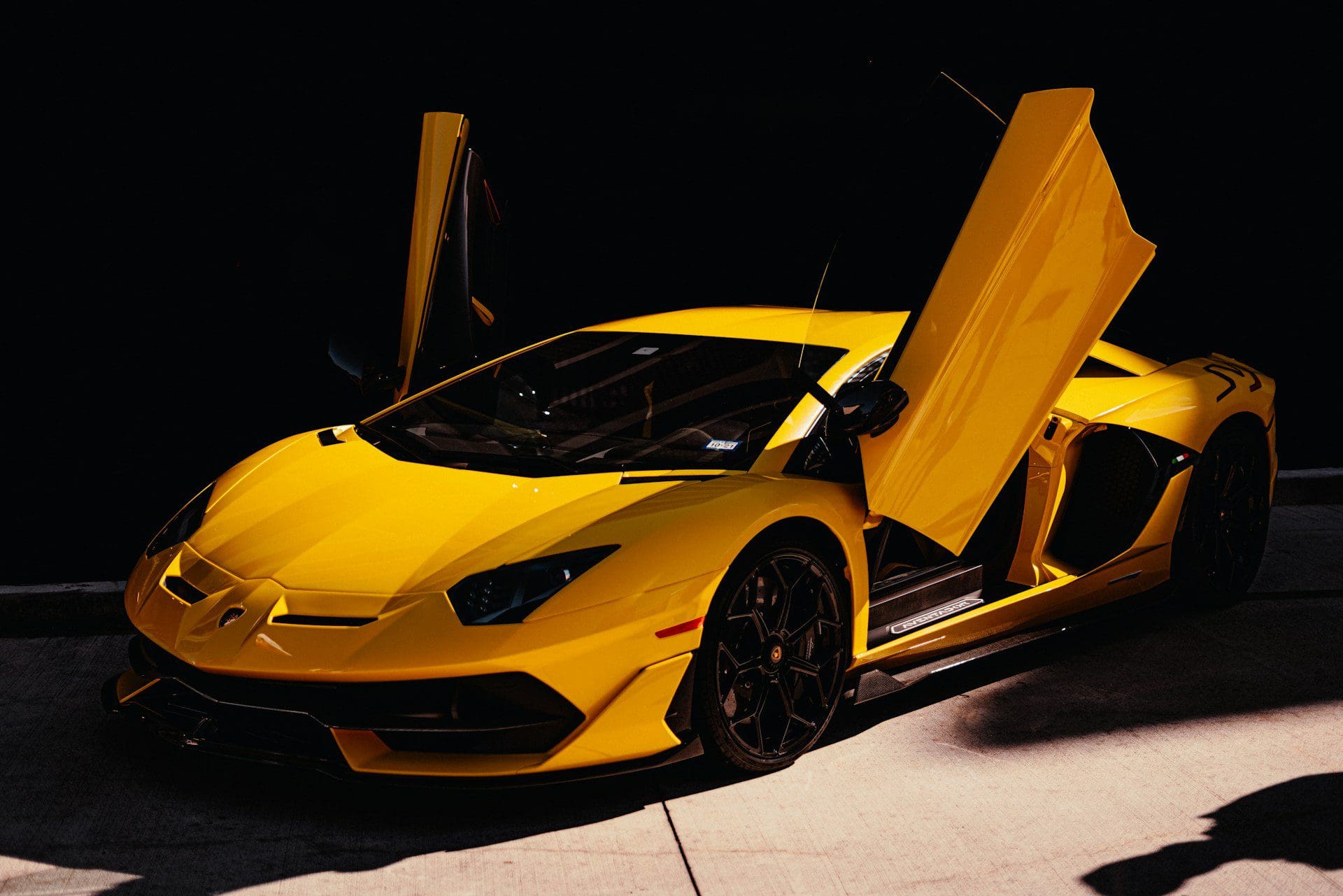 A vibrant yellow Lamborghini Aventador SVJ with its iconic scissor doors open, parked in a dimly lit setting.