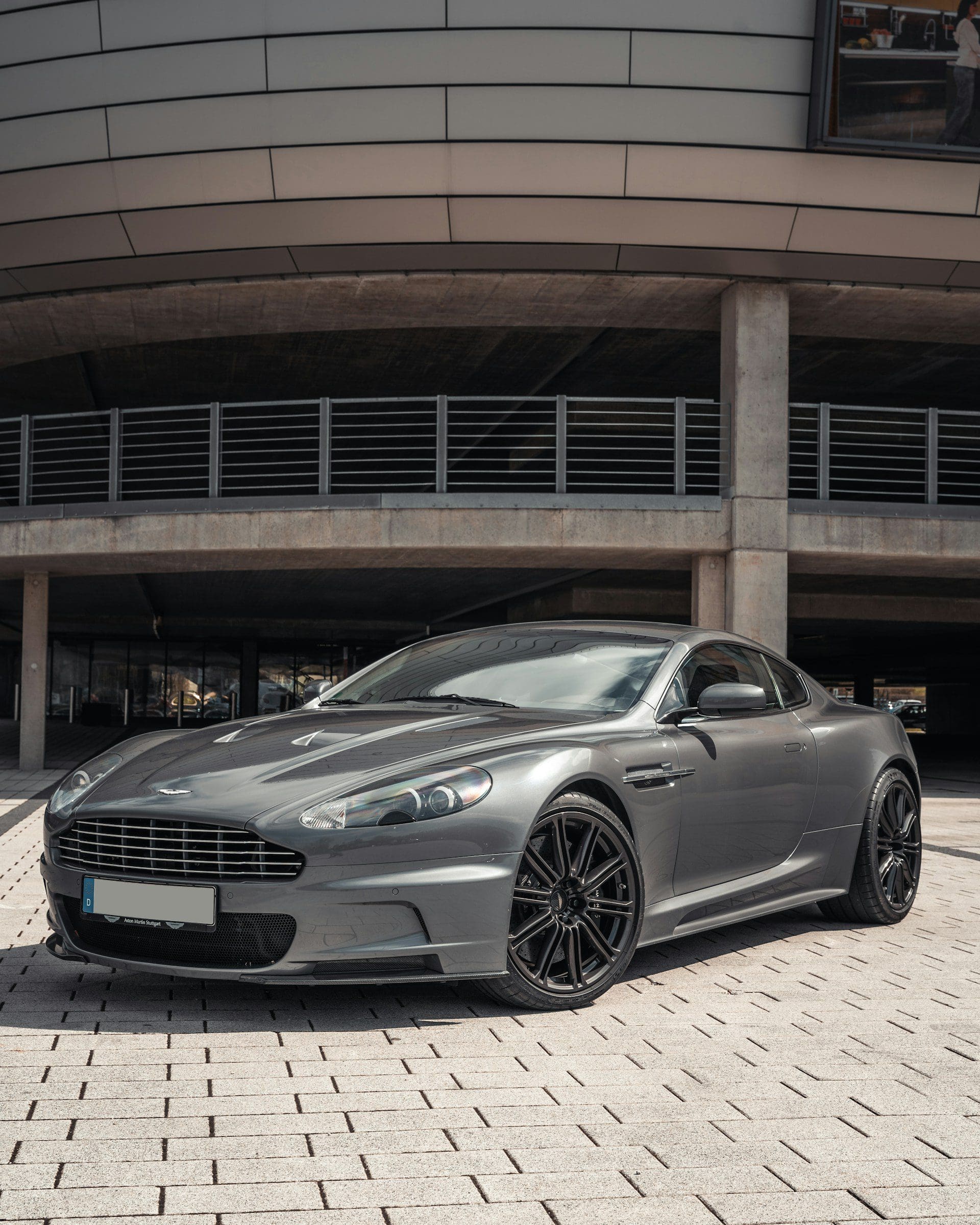 A sleek, dark gray Aston Martin DBS parked on a paved surface in front of a modern concrete building.
