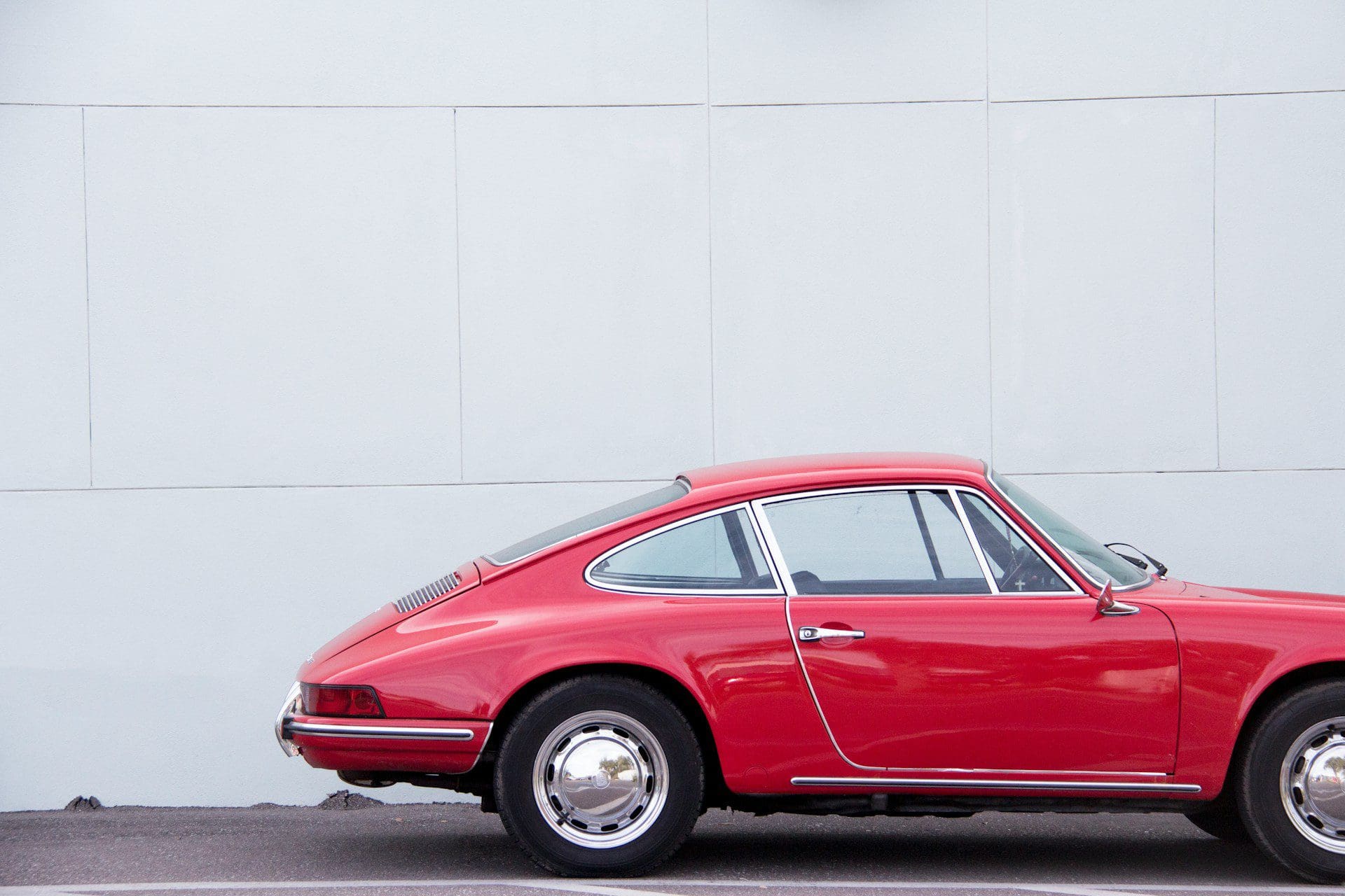 A side view of a classic red Porsche 911 parked in front of a light gray wall, highlighting its vintage design and chrome accents.