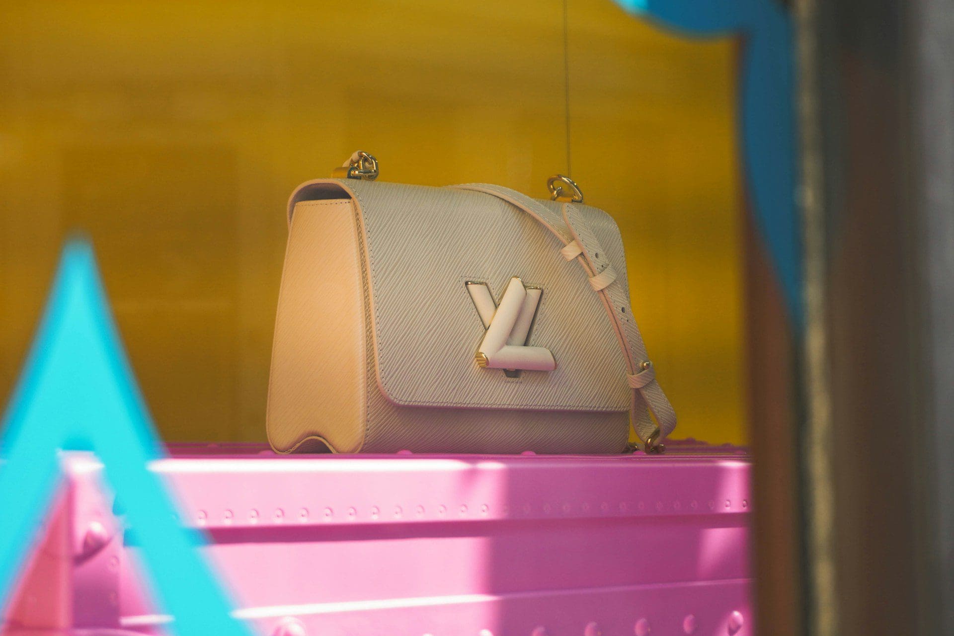 A light pink designer handbag with a gold logo sits on a bright pink display shelf, visible through a window.