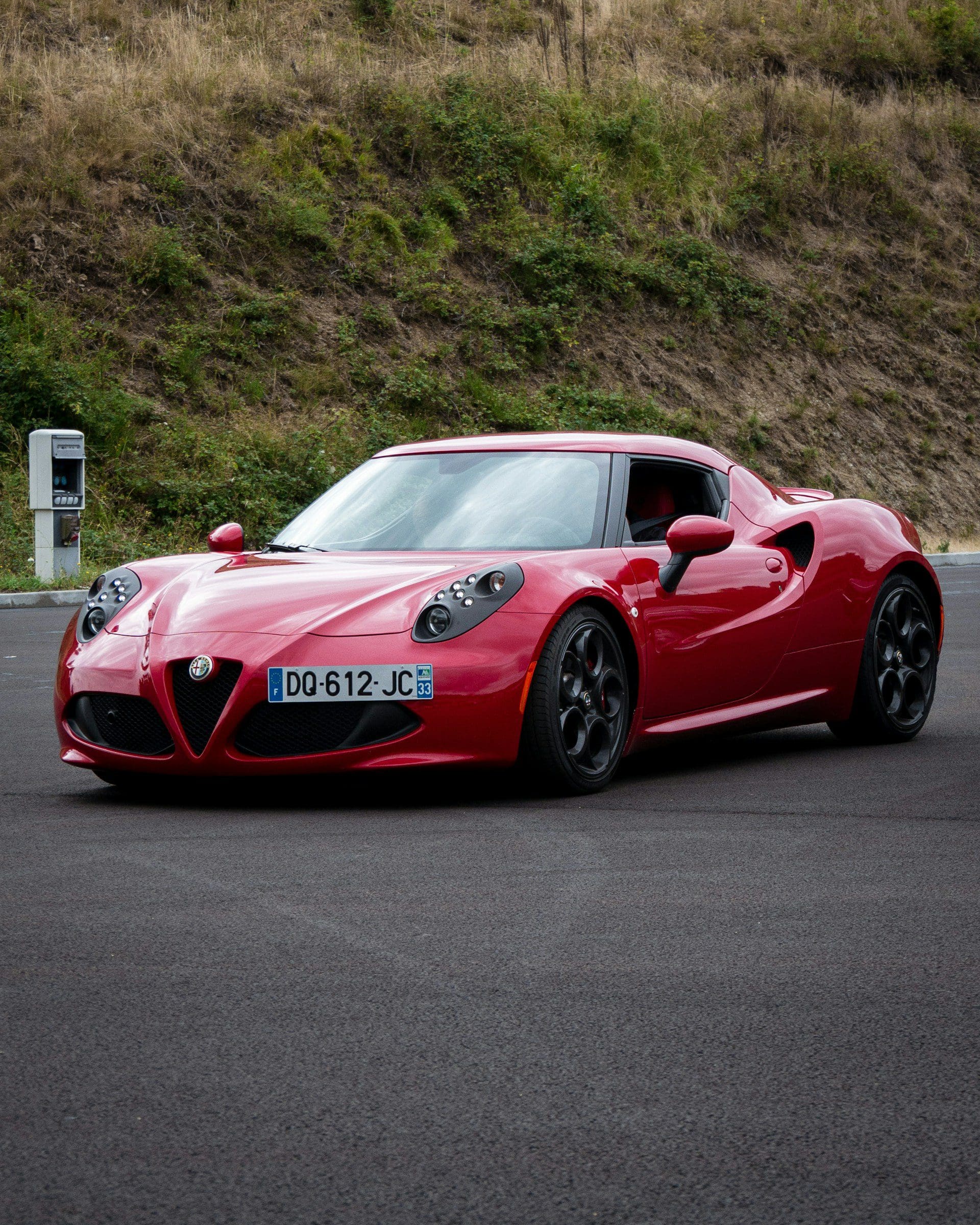 A sleek red Alfa Romeo 4C sports car is parked on asphalt with a grassy hill and charging station in the background.