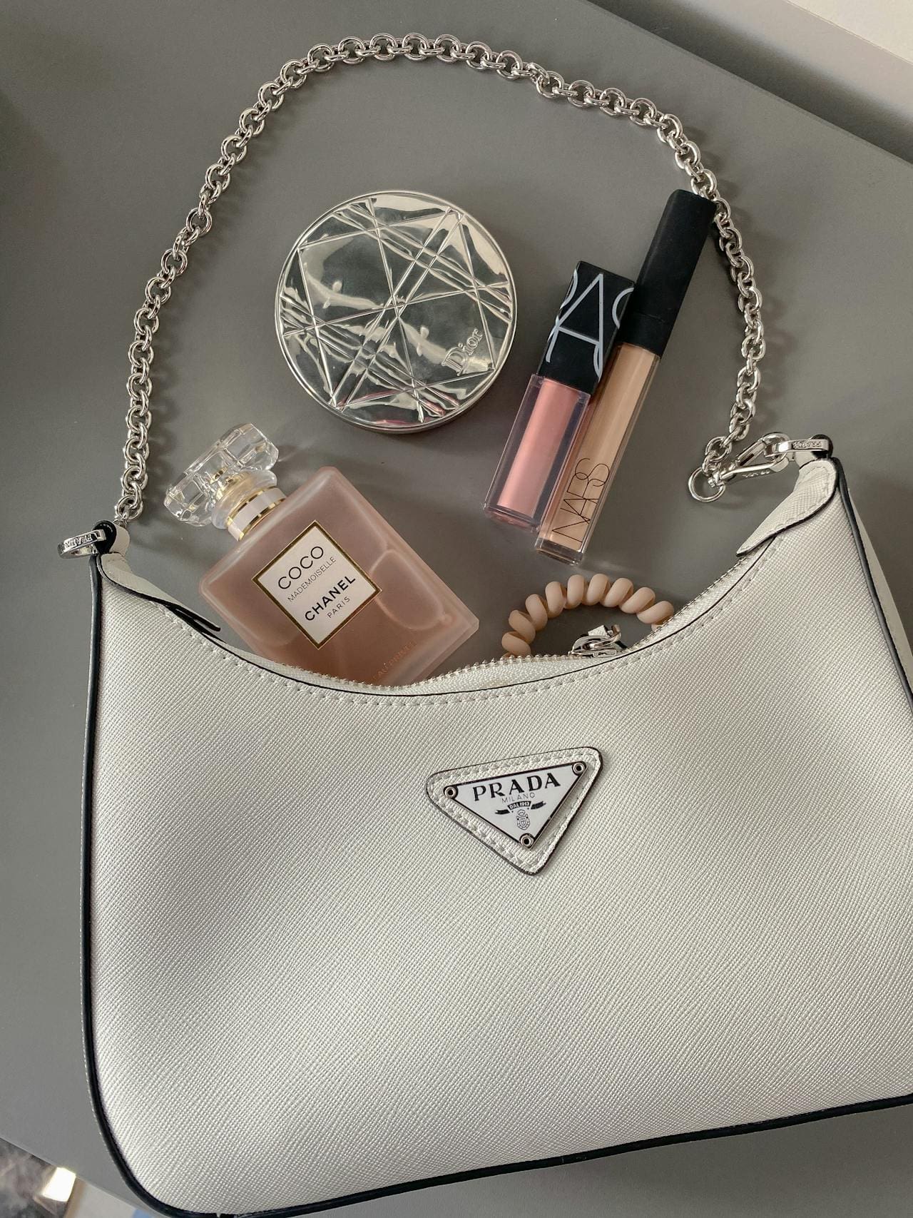 A Prada purse with Chanel perfume, NARS lip gloss, and a compact on a grey surface.