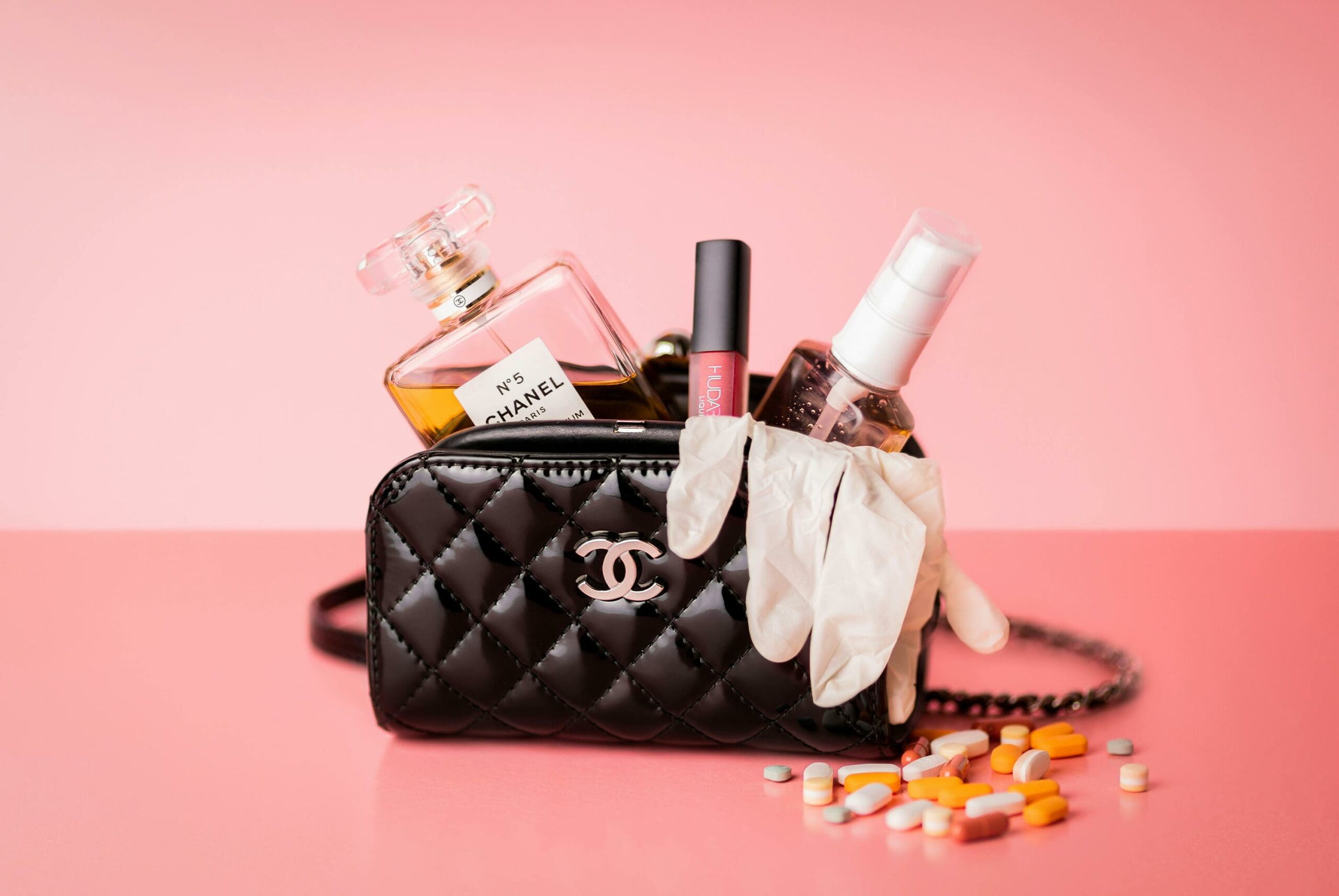 A black Chanel handbag filled with perfume, lip gloss, hand sanitizer, and medical gloves, with pills scattered around it.