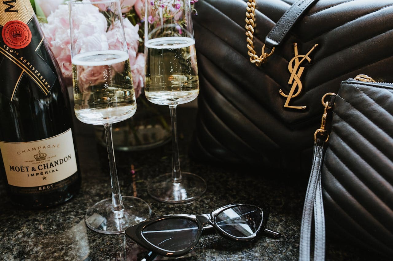 A bottle of Moët & Chandon champagne, two glasses of champagne, a black quilted handbag, and sunglasses.
