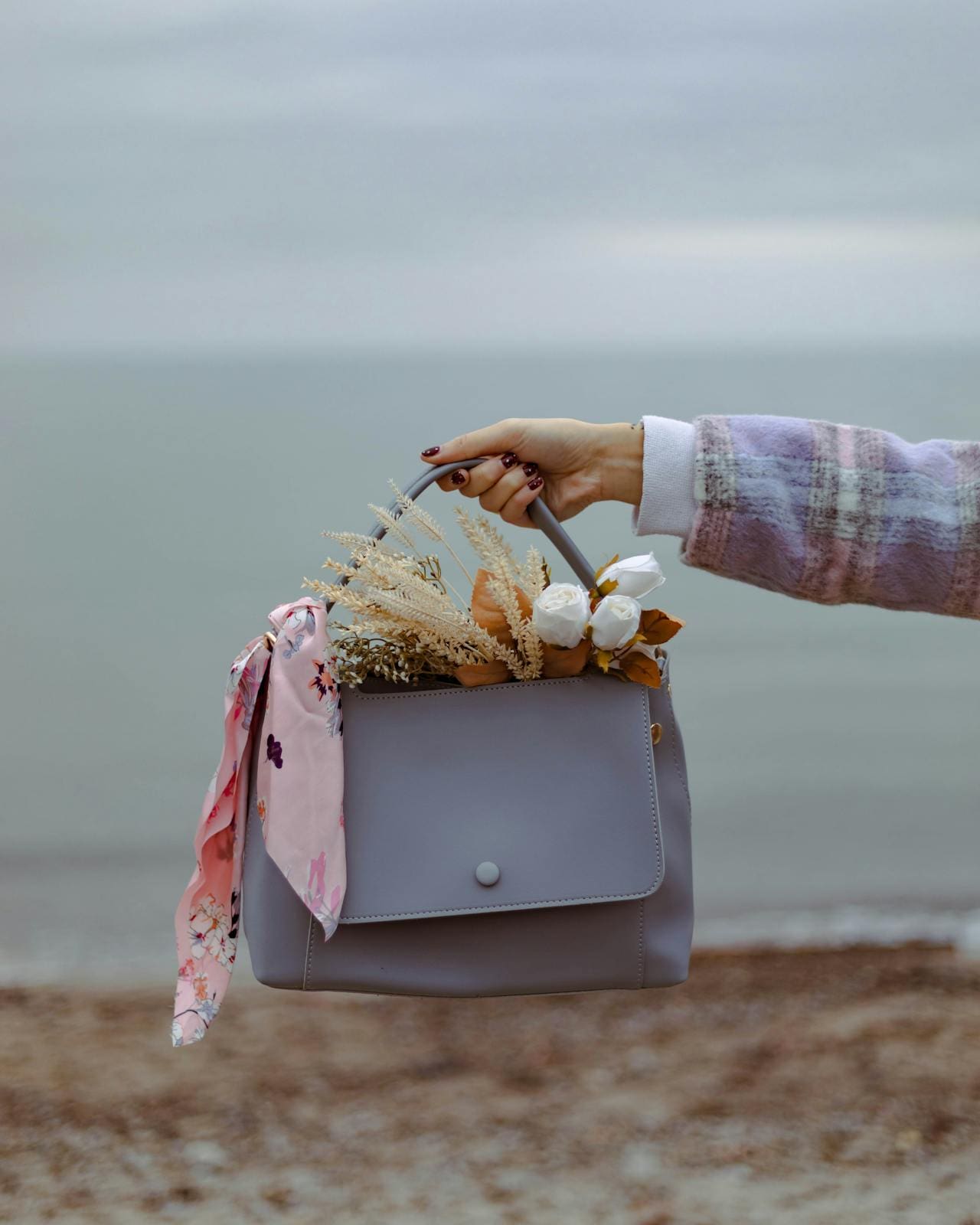 A person holds a grey handbag filled with flowers and a pink scarf against a blurry background of the sea.