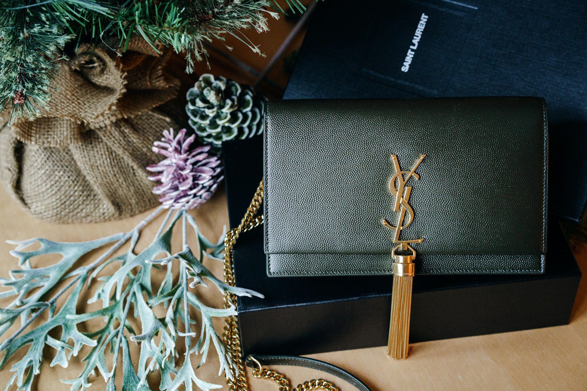 A dark green Yves Saint Laurent clutch with a gold YSL logo and tassel, surrounded by natural decorative elements.