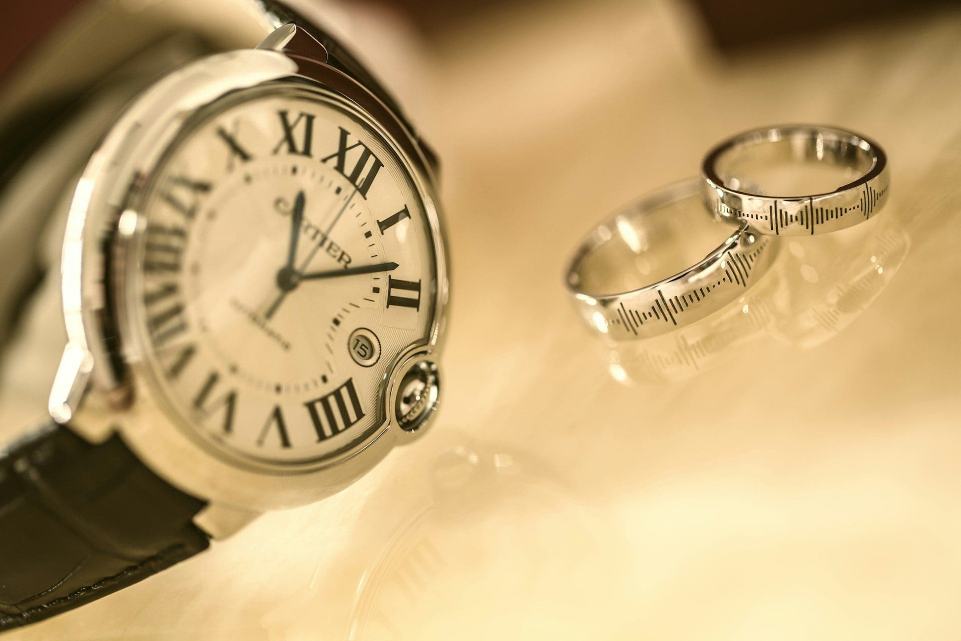 A close-up of a silver Cartier watch with Roman numerals next to two silver wedding bands with soundwave engravings.