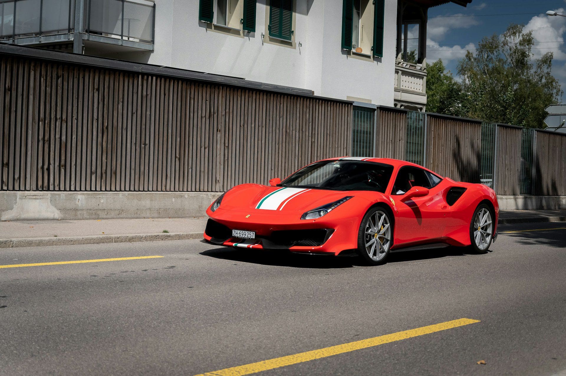 A red Ferrari 488 Pista with white and green racing stripes drives on a sunny street.